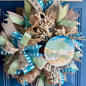 May include: A decorative wreath featuring tan mesh, light green and striped ribbons, and a round sign with a sea turtle illustration and the word Welcome. The wreath is accented with white coral-like sprigs and a teal starfish on a blue door.