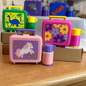 May include: A collection of miniature plastic lunch boxes and matching thermoses in various colors. The lunch boxes feature graphic prints including a blue dinosaur, a pink unicorn, and colorful flowers. Each set is connected by a small metal chain.