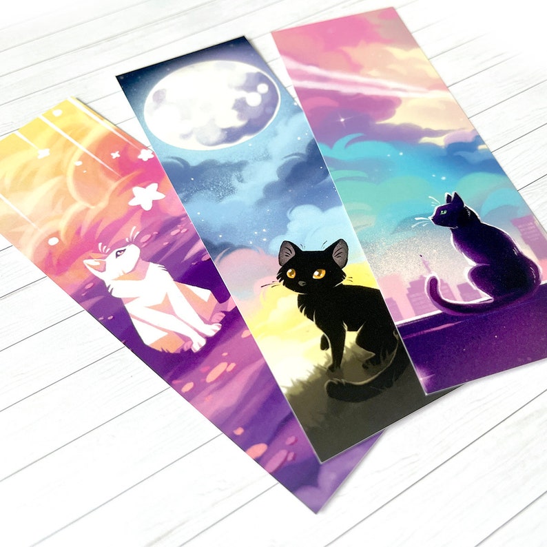 Op de afbeelding: Drie ge&iuml;llustreerde bladwijzers met katten. E&eacute;n toont een witte kat onder een sterrenhemel, een andere een zwarte kat onder een volle maan en de derde een zwarte kat op een dak met een skyline. De achtergronden hebben kleurverlopen.