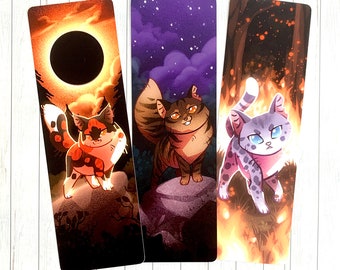 May include: Three illustrated bookmarks featuring cats in different poses. The first bookmark features a black and orange cat with a dark background. The second bookmark features a brown and white cat with a purple background. The third bookmark features a gray and white cat with a fiery orange background.