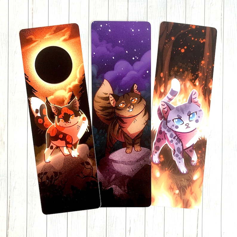 May include: Three illustrated bookmarks featuring cats in different poses. The first bookmark features a black and orange cat with a dark background. The second bookmark features a brown and white cat with a purple background. The third bookmark features a gray and white cat with a fiery orange background.