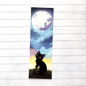 Op de afbeelding: Bladwijzer met een illustratie van een zwarte kat die naar een volle maan in een nachtelijke hemel kijkt. De illustratie bevat een kleurengradi&euml;nt, met een gele en roze zonsondergang aan de onderkant en een sterrenhemel erboven.