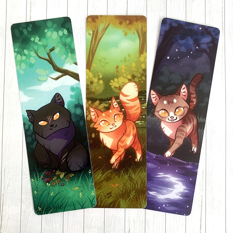 May include: Three bookmarks featuring cartoon cats in different settings. The first bookmark shows a black cat with green eyes in a forest setting. The second bookmark shows an orange cat with green eyes in a forest setting. The third bookmark shows a tabby cat with green eyes in a night sky setting.