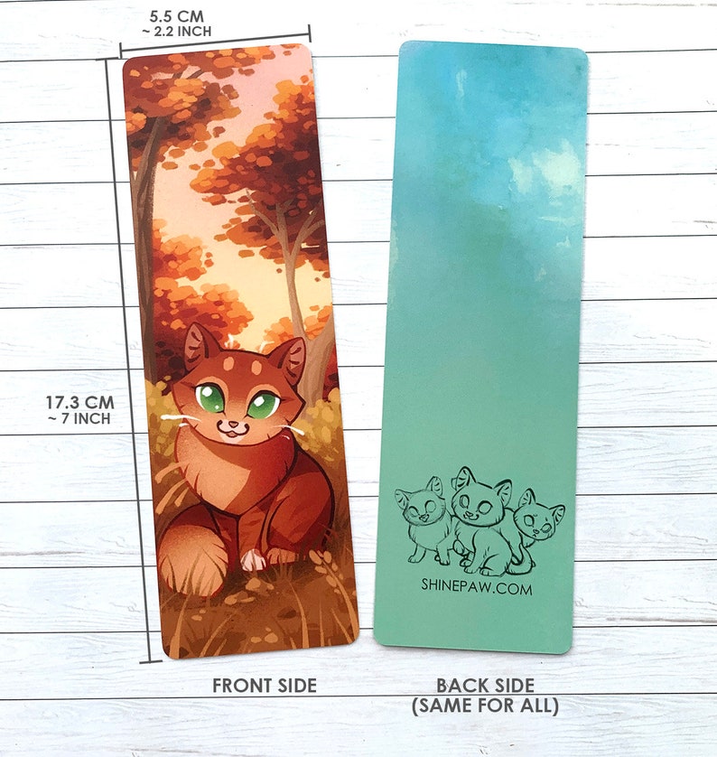 May include: A bookmark featuring a cartoon orange tabby cat sitting in a forest with fall foliage. The back side of the bookmark has a light blue background with three cartoon cats in black outline. The bookmark is 7 inches tall and 2.2 inches wide. The text "SHINEPAW.COM" is printed on the back of the bookmark.