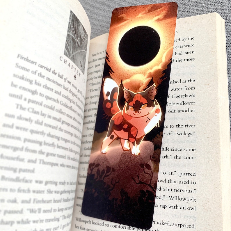 May include: A bookmark featuring a calico cat with orange, black, and white fur standing on a rock. The cat is looking to the right. The background is a sunset with a large black circle in the middle. The text "Chapter 15" is visible in the upper left corner.