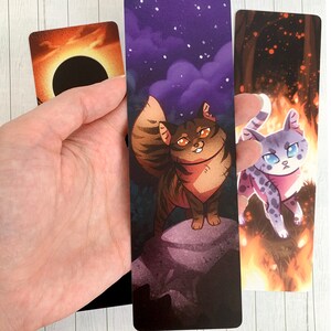 May include: Three bookmarks featuring illustrations of cats. The first bookmark has a dark orange background with a black circle in the center. The second bookmark has a purple background with a brown tabby cat standing on a rock. The third bookmark has a dark orange background with a white and gray cat standing in flames.