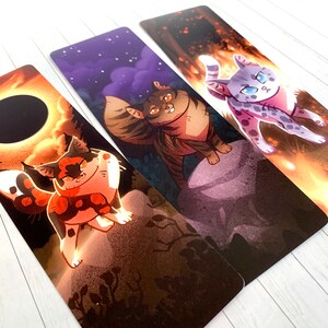 May include: Three bookmarks featuring colorful illustrations of cats. The first bookmark has a black and red cat with a white chest and paws, standing on a rock with a dark background. The second bookmark has a brown tabby cat with a white chest and paws, standing on a rock with a purple and blue background. The third bookmark has a white and gray cat with black spots, standing on a rock with a fiery orange background.