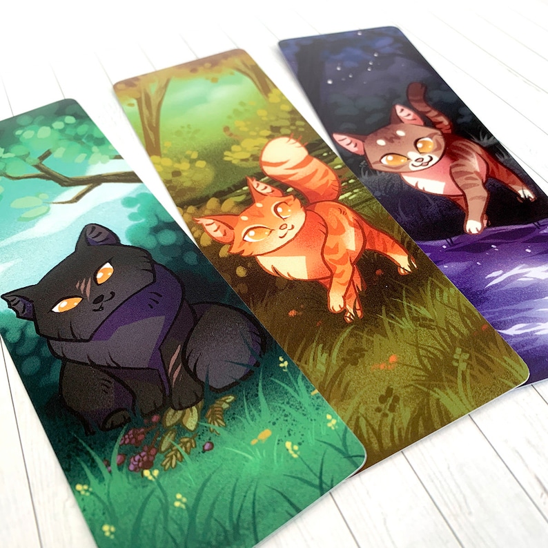 May include: Three illustrated bookmarks featuring cats in different settings. The first bookmark shows a black cat sitting in a green grassy field. The second bookmark shows an orange tabby cat sitting in a green forest. The third bookmark shows a gray tabby cat walking on a purple and blue river.