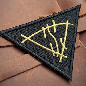 May include: A triangular black embroidered patch featuring a gold-colored stitched symbol. The patch is attached to a brown leather surface with a visible strap. The design consists of intersecting lines forming a geometric shape with vertical marks.