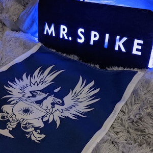 Op de afbeelding: Een blauwe stoffen vlag met een wit gestileerd adelaarontwerp. De vlag ligt op een wit pluche oppervlak. Een zwart bord met witte letters leest "MR. SPIKE" in hoofdletters.