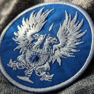 May include: A round, embroidered patch featuring a detailed white eagle design on a blue background. The patch has a white border and is set against a gray, textured fabric. The eagle design includes outstretched wings and a crest.
