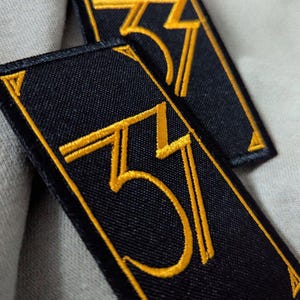 May include: Two rectangular black embroidered patches with the number "37" in gold thread. The patches have a gold border and are set against a light gray fabric background.