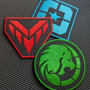 Puede incluir: Tres parches bordados con diseños distintos: un emblema rojo y negro en forma de escudo, un parche cuadrado azul y negro con una estrella central y un parche circular verde y negro que representa el perfil de una cabeza de león estilizada.