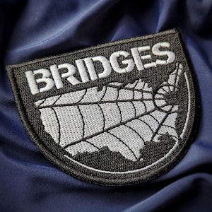 May include: A navy blue patch with the word "BRIDGES" in white, arched above a stylized map of the United States. The map is in white with a web-like design, all set against a black border.
