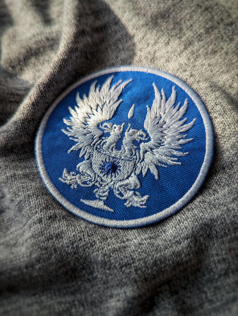 May include: A round, embroidered patch featuring a detailed white emblem on a blue background. The emblem depicts a double-headed eagle with outstretched wings, surrounded by a white border. The patch is set against a gray fabric.