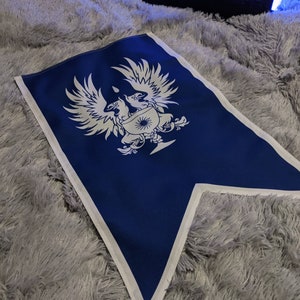 May include: A blue fabric flag with a white design of two birds with a shield and a sun in the center. The flag is on a gray fuzzy surface. The text "MR. SPIKE" is visible in the background.
