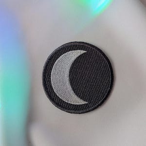 K&ouml;nnte beinhalten: Ein runder, schwarzer, gestickter Aufn&auml;her mit einem Halbmond-Design in Hellgrau. Der Aufn&auml;her hat einen schwarzen Rand und ist vor einem wei&szlig;en und schillernden Hintergrund platziert.