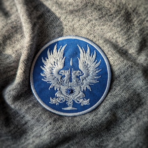 May include: A round, blue patch with a detailed white embroidered design featuring a crest with two winged creatures. The patch is set against a gray, textured fabric background.