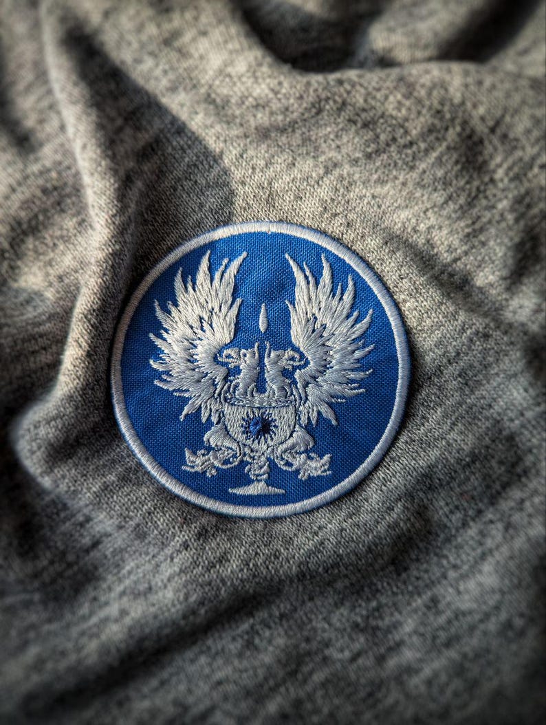 May include: A round, blue patch with a detailed white embroidered design featuring a crest with two winged creatures. The patch is set against a gray, textured fabric background.