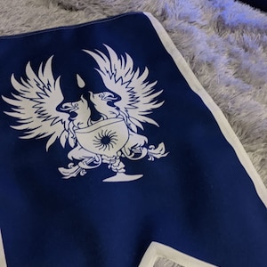 May include: A blue fabric flag with a white design of two stylized birds flanking a goblet with a sun inside. The birds have wings spread out and are facing each other.