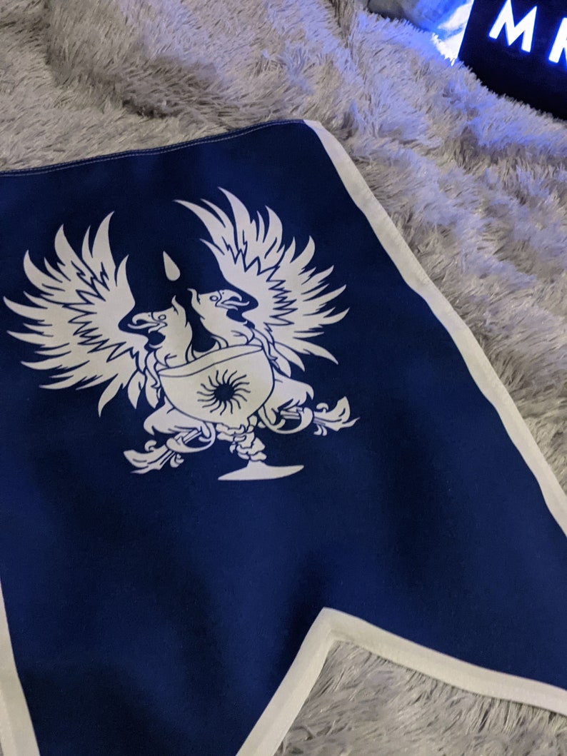 May include: A blue fabric flag with a white design of two stylized birds flanking a goblet with a sun inside. The birds have wings spread out and are facing each other.