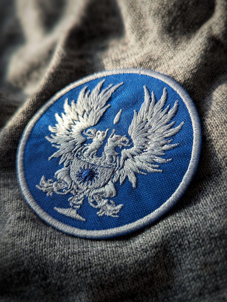 May include: A round, embroidered patch featuring a detailed white eagle design on a blue background. The patch has a white border and is set against a gray, textured fabric. The eagle design includes outstretched wings and a crest.