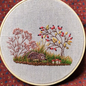 May include: Embroidered hoop art featuring a brown hedgehog, green grass, red berries, and yellow leaves on a beige fabric background.