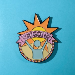 May include: A yellow sun with a pink banner that says "You Got Up" and a blue stick figure with arms raised in the air. The sun and stick figure are on a yellow background with a pink and orange border.