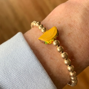 Pode incluir: Um bracelete de contas douradas com um amuleto de taco amarelo. O bracelete está no pulso de uma pessoa.
