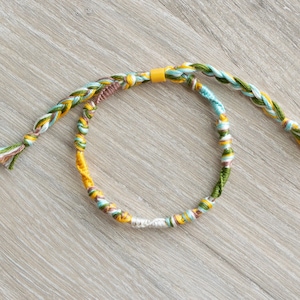 May include: A braided friendship bracelet with yellow, green and blue threads. The bracelet is tied with a small yellow bead.