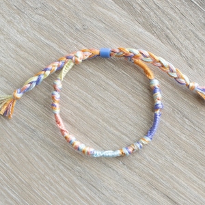 May include: A multi-colored braided friendship bracelet with a blue accent bead. The bracelet is made with a variety of colors, including orange, yellow, and blue.