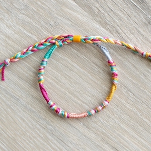 May include: A multi-colored friendship bracelet with a braided design and a yellow accent. The bracelet is made of thin threads in shades of pink, teal, white, and gold.