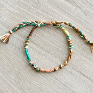 May include: A braided friendship bracelet with brown, beige, and teal threads. The bracelet has a knotted closure and tassels on each end.