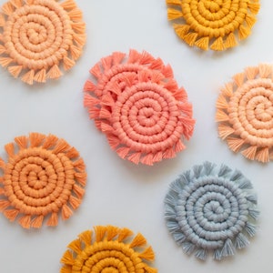 May include: A set of six colorful crocheted coasters with a spiral design and fringe. The coasters are in shades of blue, orange, and yellow.