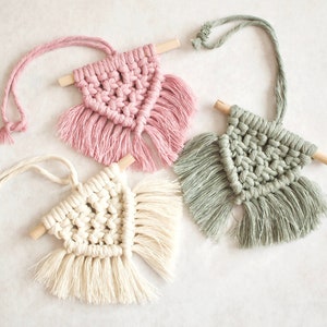May include: Three macrame wall hangings with wooden dowels and fringe. The hangings are in pink, white, and green.