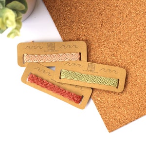 Könnte beinhalten: Drei gewebte Freundschaftsarmbänder in Korallen-, Pfirsich- und Grüntönen, präsentiert auf Kartonverpackungen. Die Verpackung trägt den Text "Oasis Strings" und ein Wellenmuster. Die Armbänder befinden sich auf einem weißen und Kork-Hintergrund.