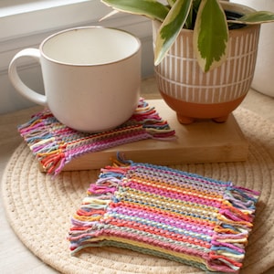 May include: Two rectangular woven coasters featuring a multi-colored striped pattern in shades of pink, blue, yellow, orange, and green. Each coaster has a textured surface with short, knotted fringe detailing along the two shorter edges.