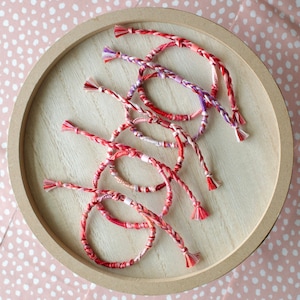 May include: A collection of colorful friendship bracelets made with braided string in shades of pink, red, and white. The bracelets have tassels at the ends and are arranged on a wooden surface.