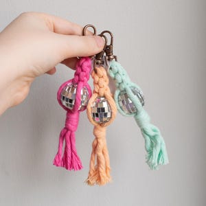 May include: Three disco ball keyrings in pink, peach, and mint green. Each keyring features a small, mirrored disco ball attached to a knotted cord and a bronze-coloured clasp. The keyrings are held in a hand against a neutral background.