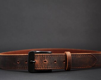 May include: A brown leather belt with a black buckle. The belt has a distressed finish and visible stitching. The buckle is rectangular and the belt is laid flat against a dark background.
