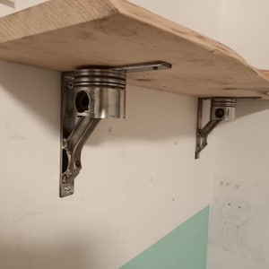 Könnte beinhalten: Ein Holzregal mit einer lebenden Kante wird von zwei Metallkonsolen getragen. Die Konsolen sind aus wiederverwendeten Autoteilen hergestellt, darunter ein Kolben. Das Regal ist an einer weißen Wand montiert.