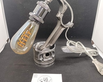 Puede incluir: Una lámpara de escritorio única hecha de piezas de motor recicladas. La lámpara presenta un portalámparas plateado, una bombilla Edison transparente con filamento naranja y una base de pistón. El marco está hecho de una biela. Un cable de alimentación blanco está conectado.