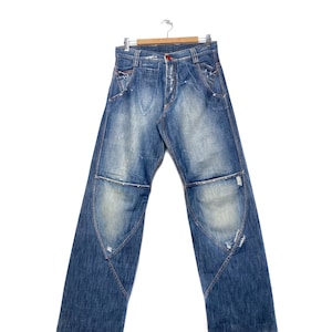 Può includere: Un paio di jeans blu in denim con dettagli usurati e rinforzi alle ginocchia. I jeans hanno una vestibilità rilassata e una gamba dritta.