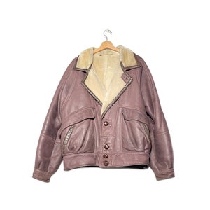 Può includere: Una giacca di pelle marrone con colletto in montone e chiusura con bottoni marroni. La giacca ha due grandi tasche sul davanti e una tasca con zip sulla manica sinistra.