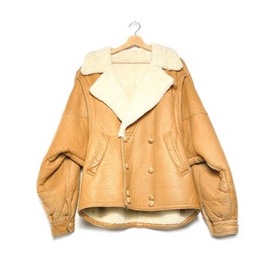 Può includere: Giacca in pelle marrone chiaro con fodera e colletto in montone color crema. La giacca doppiopetto ha quattro bottoni e due tasche anteriori. È appesa a una gruccia di legno su sfondo bianco.