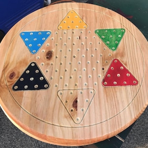 Puede incluir: Un tablero de juego de madera circular con un diseño de estrella. La estrella está formada por cinco triángulos, cada uno pintado de un color diferente: amarillo, azul, negro, rojo y verde. El tablero tiene un gran número de agujeros perforados en él.