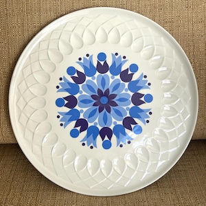 May include: White ceramic serving platter with a blue floral design in the center. The floral design is surrounded by a pattern of blue and purple tulip-like shapes. The rim features a repeating embossed design.