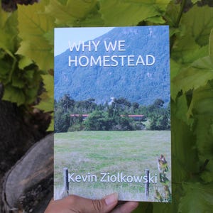 May include: A book titled "WHY WE HOMESTEAD" with a landscape image of a mountain, field, and trees. The author's name, Kevin Ziolkowski, is printed at the bottom. The book is held against a backdrop of green leaves.