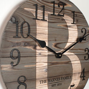 May include: A round wooden clock with black hands and black numbers. The clock face is made of reclaimed wood with a natural wood grain. The clock face has a large letter "B" in the center. The clock face also has the text "THE BARNES FAMILY EST. 2016".