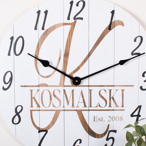 May include: White wooden clock with black Roman numeral hour markers and black hands. The clock features a large brown letter "K" with the name "KOSMALSKI" and "Est. 2008" printed below.